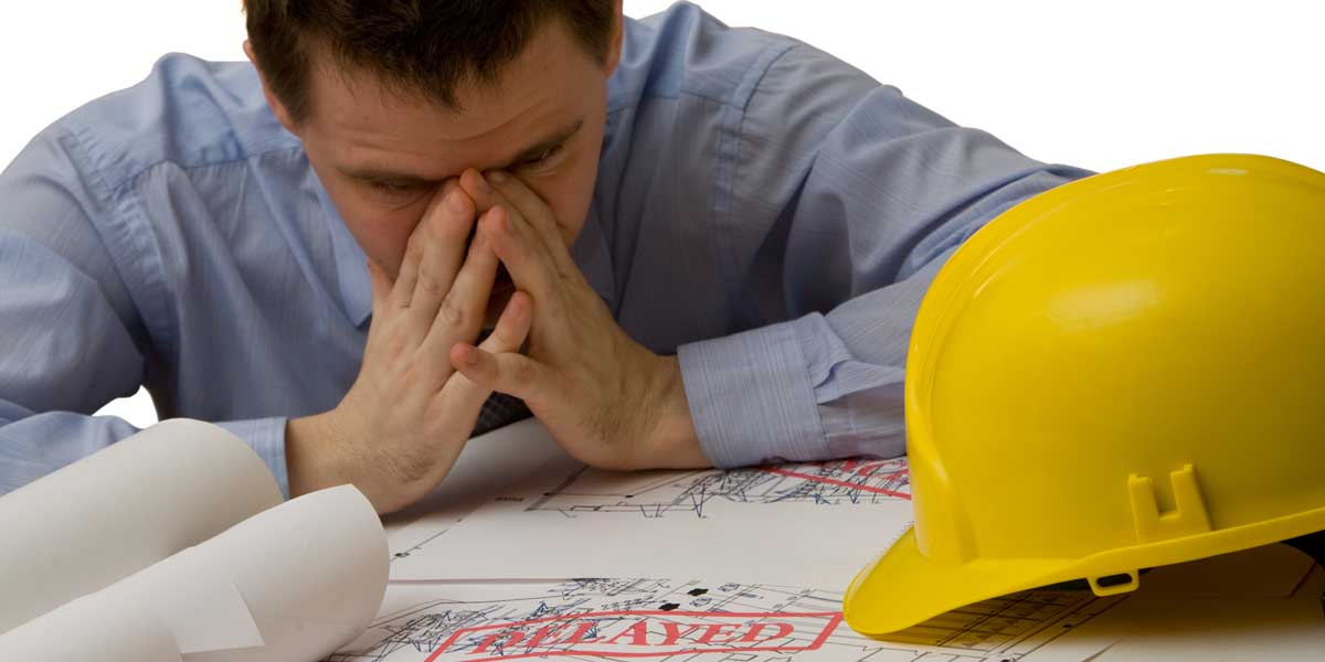 Contractor with a yellow hard hat holding his head looking at construction plans with the word delayed stamped on it
